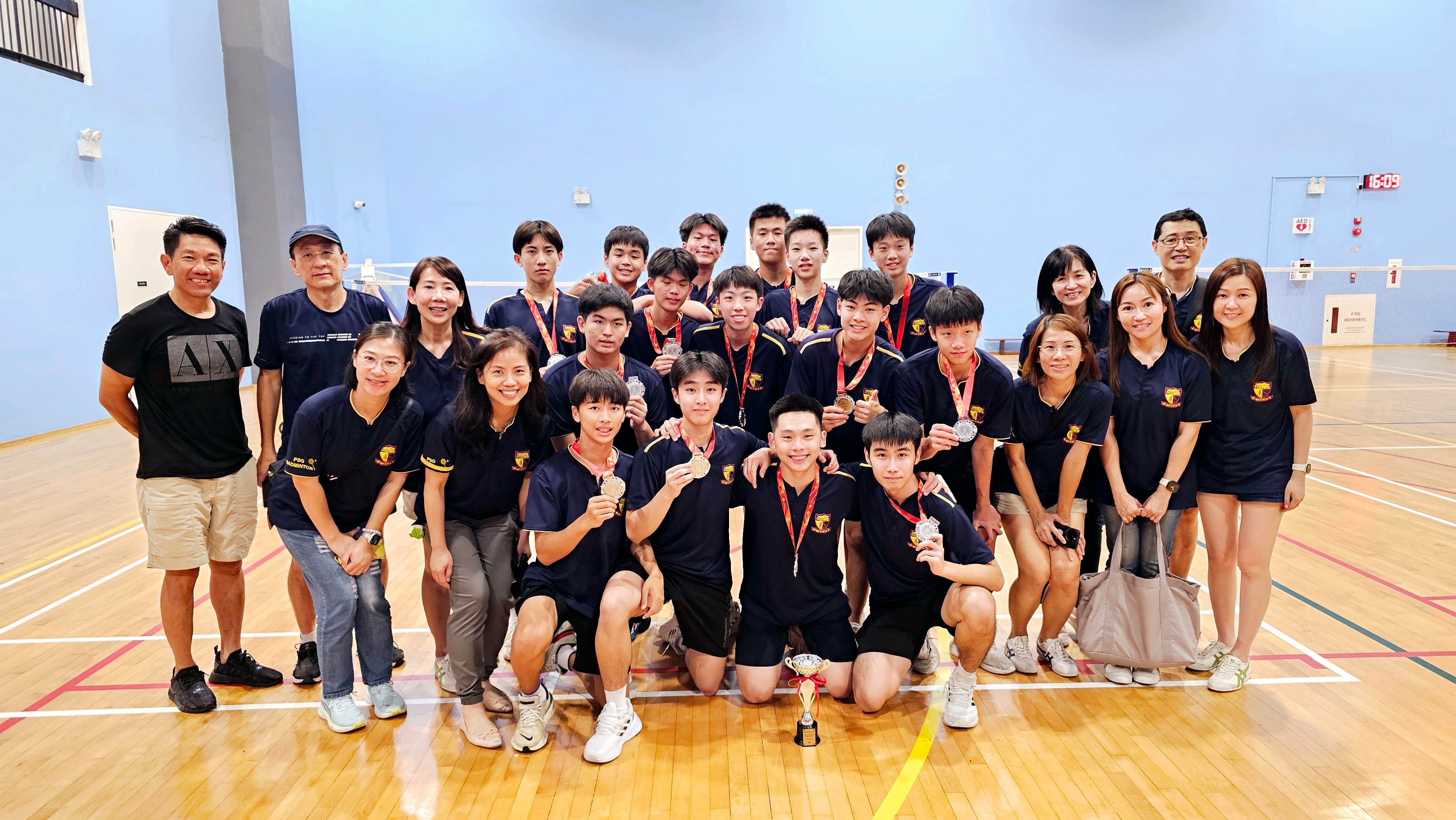 A large mixed group of students and adults in black tops poses together on a badminton court for a team photo.